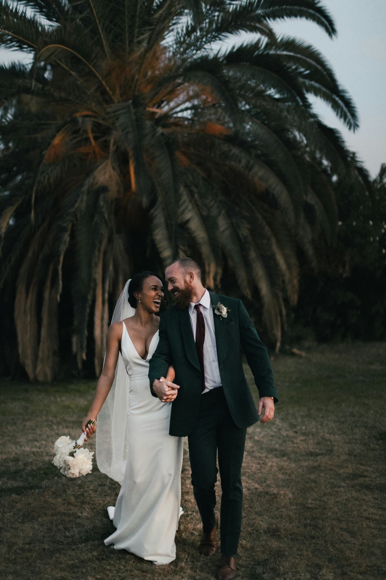 Bride and groom laughing as they walk beneath a palm at dusk
